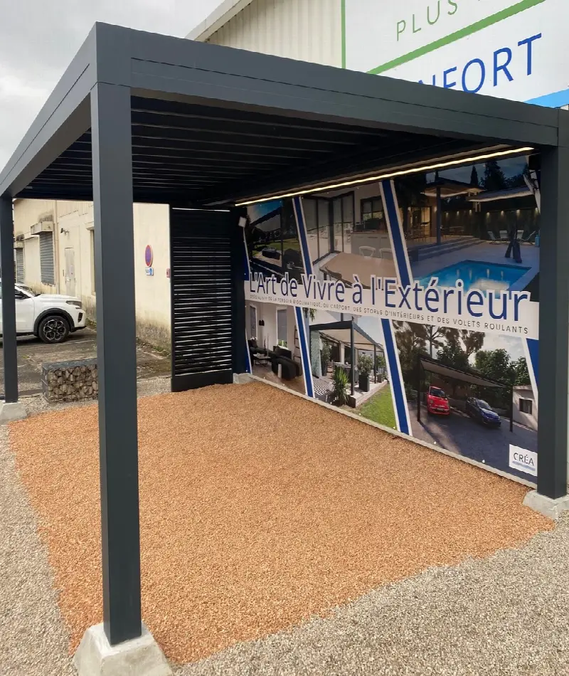 Pergola noire à lames posée sur du gravier, devant une vitrine