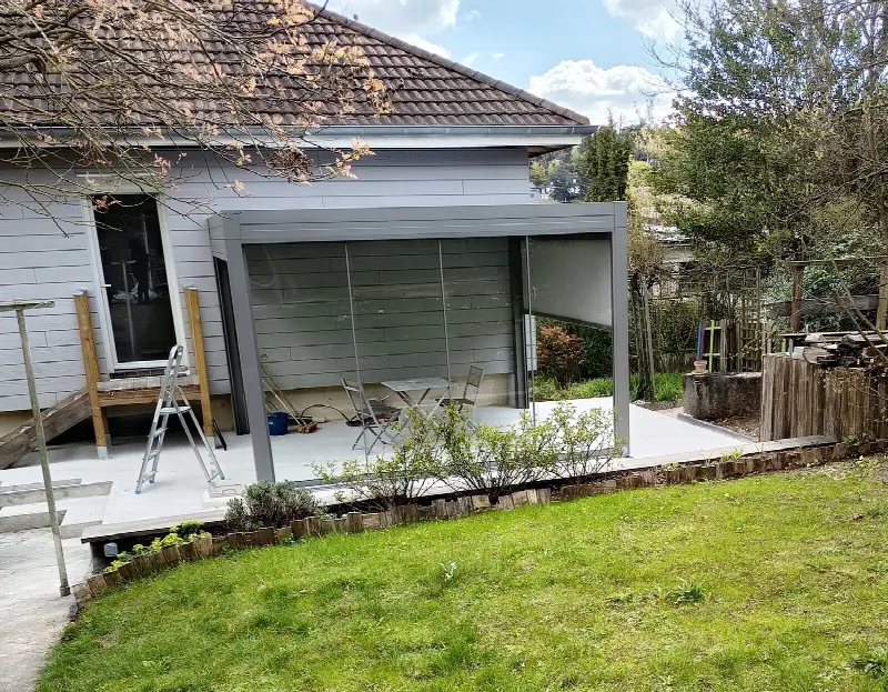 Pergola adossée à une maison à bardage blanc, store descendu, au bord d'une pelouse