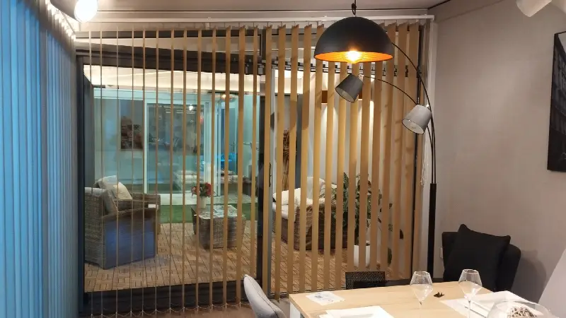 Stores à bandes verticales ocre entrouverts, laissant voir une véranda meublée d’un salon en résine tressée